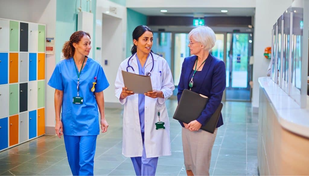 Three healthcare professionals, including a nurse, a doctor, and an administrator, walk together down a hospital corridor, talking and holding documents.