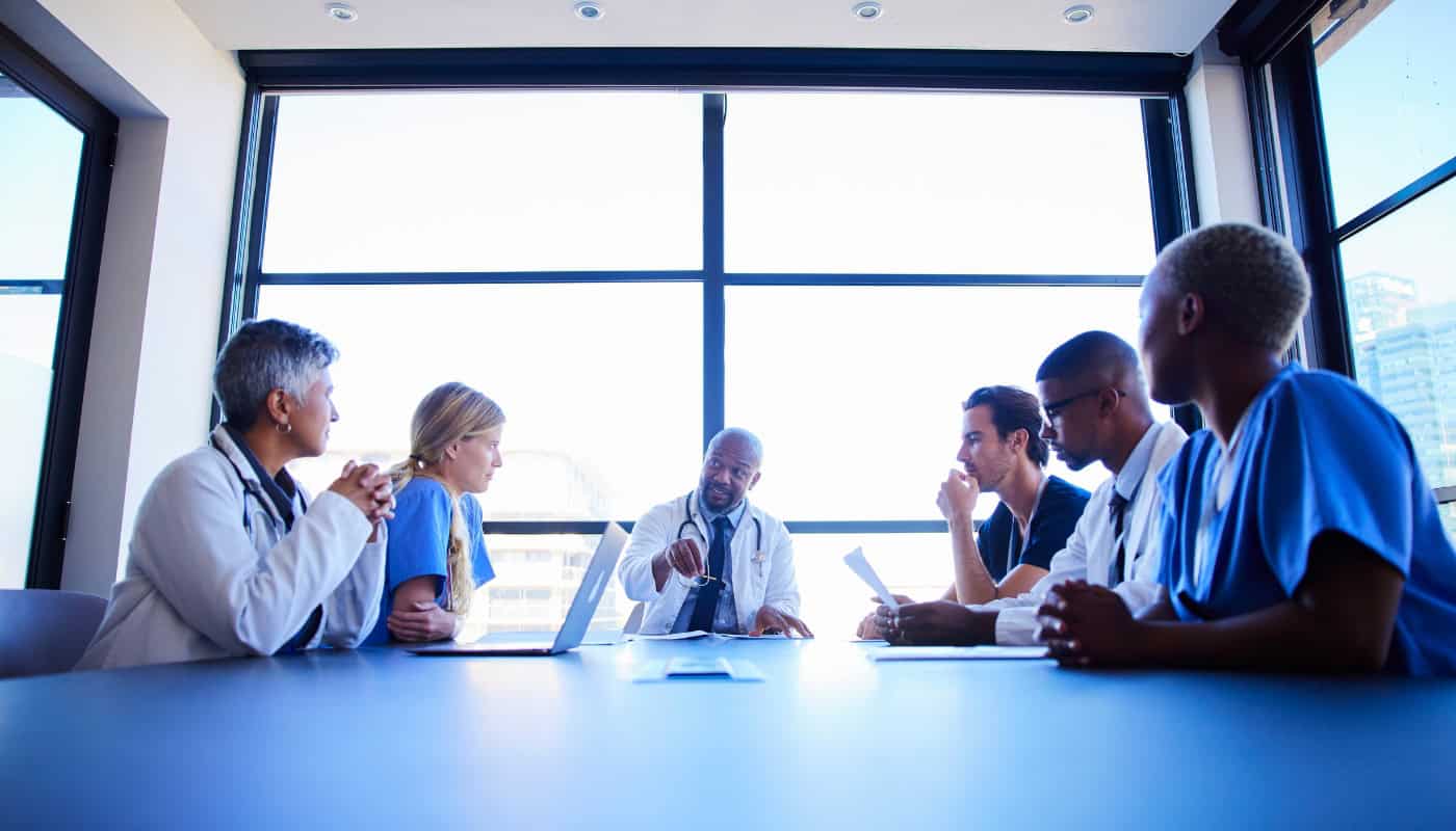 Six healthcare professionals sit around a conference table in a modern office, engaged in discussion, with papers and a laptop in front of them.