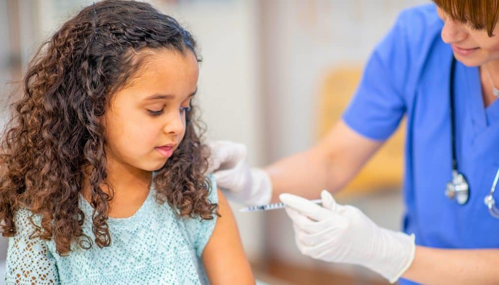 A healthcare worker in scrubs administers a vaccine to a young girl with curly hair, who looks away as she receives the injection in her upper arm.