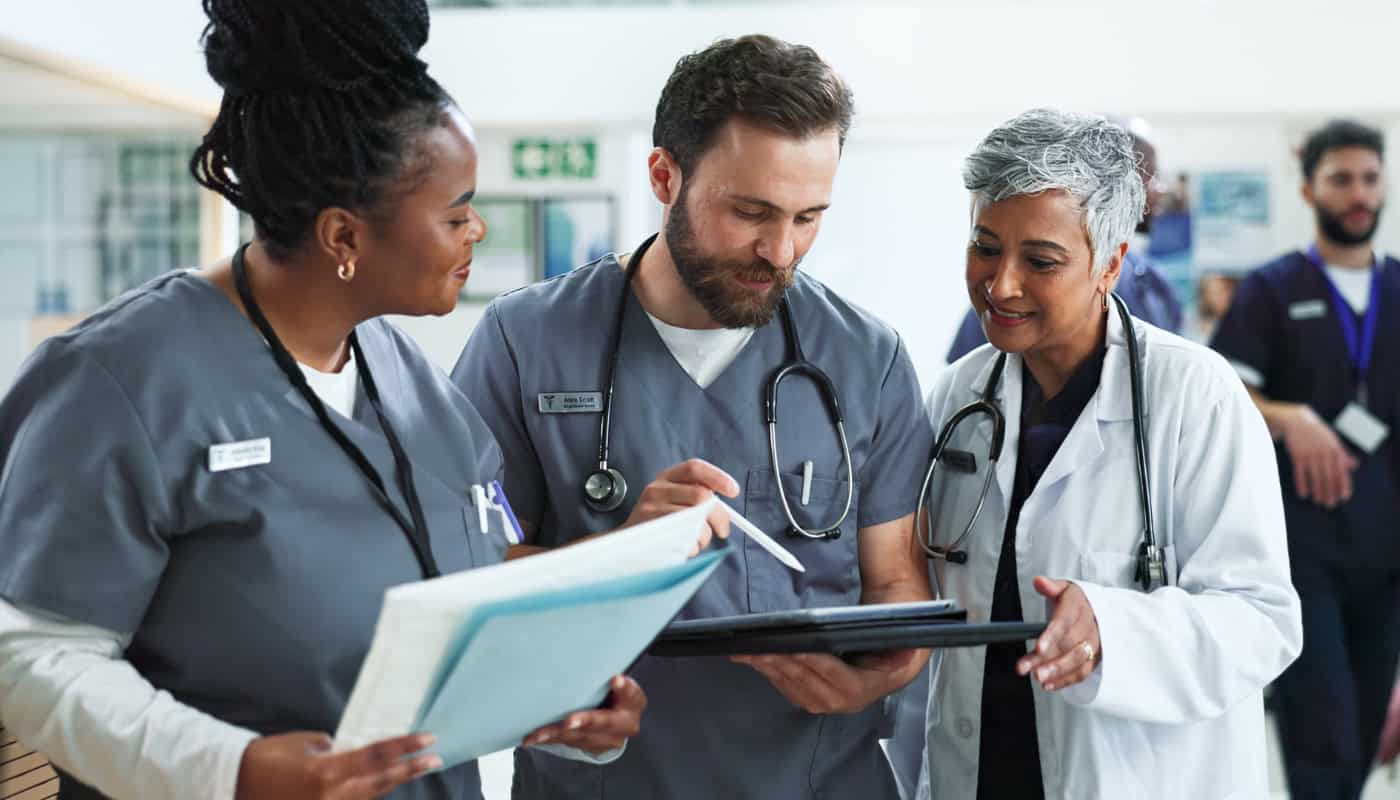 Three healthcare professionals in scrubs and lab coats confer over a digital tablet in a hospital hallway.