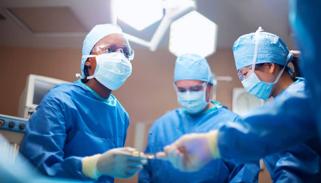 Three surgeons in blue scrubs, masks, and protective eyewear working in an operating room.