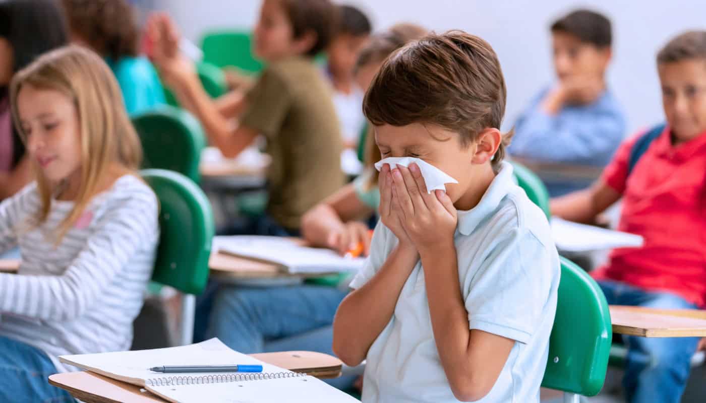 A young boy sitting at a school desk blows his nose into a tissue, with other students visible in the background.