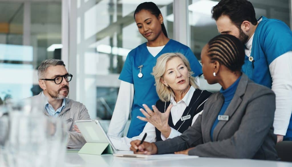 A diverse group of healthcare professionals and administrators in a meeting, with a woman gesturing while discussing a tablet.
