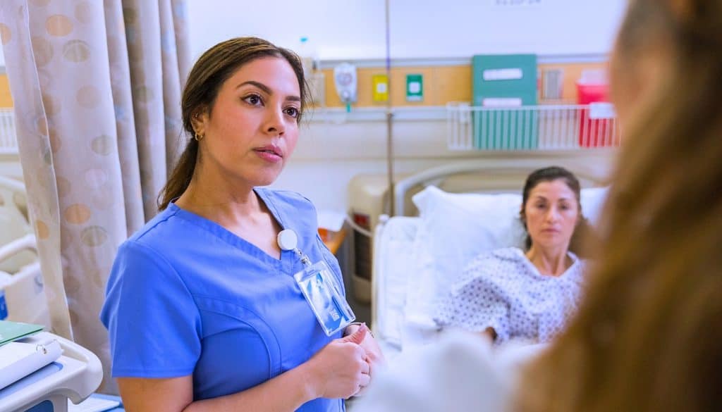 A nurse in blue scrubs speaks to a patient resting in a hospital bed.