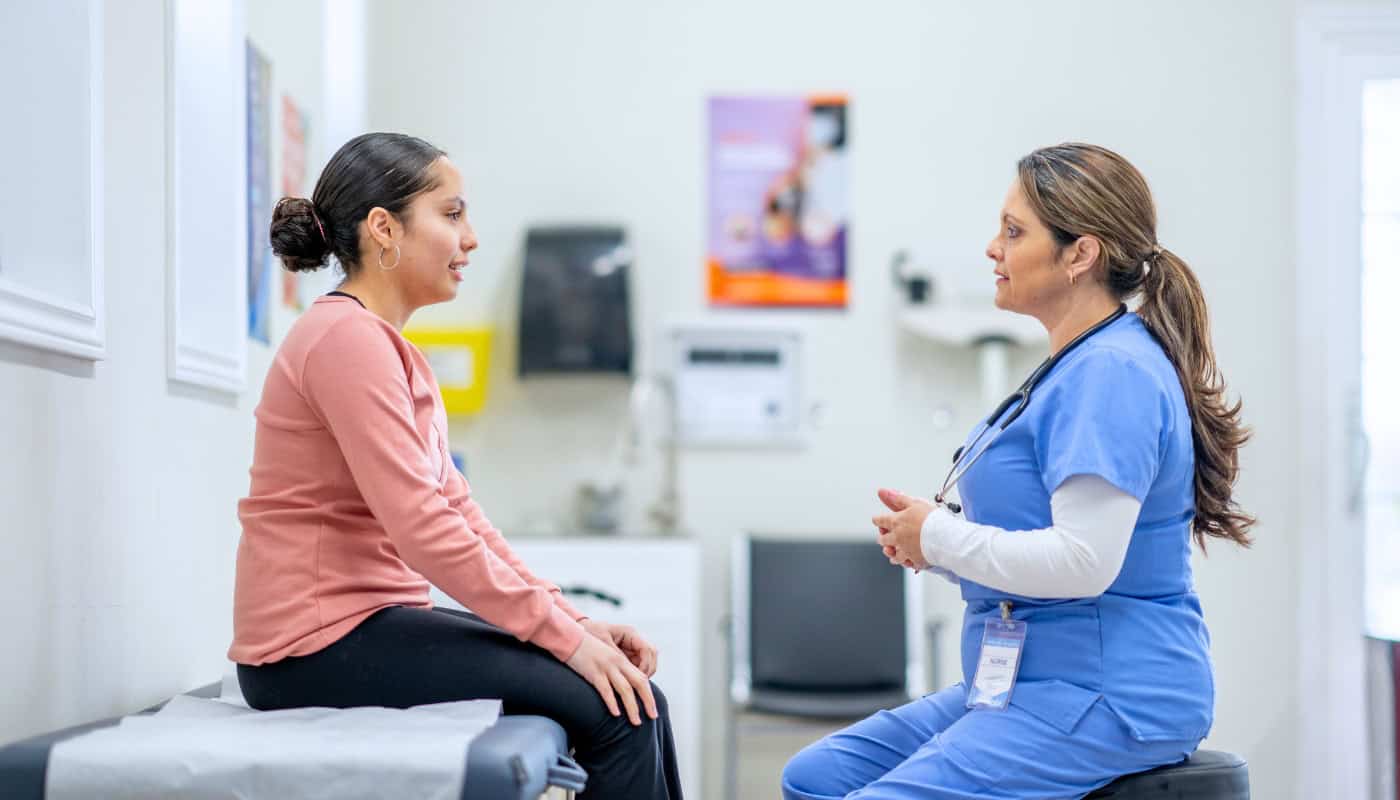 A nurse in blue scrubs talking to a patient sitting on an exam table in a clinic.