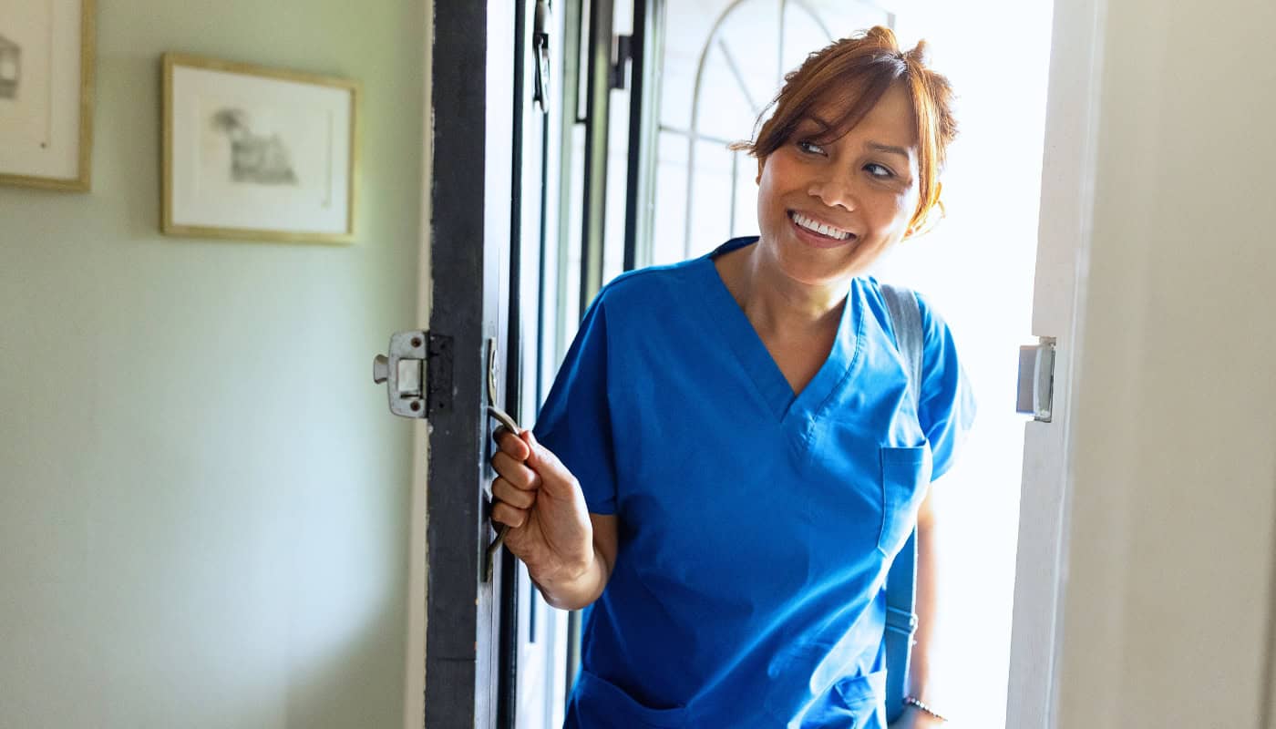 A smiling healthcare worker in blue scrubs opens a front door.