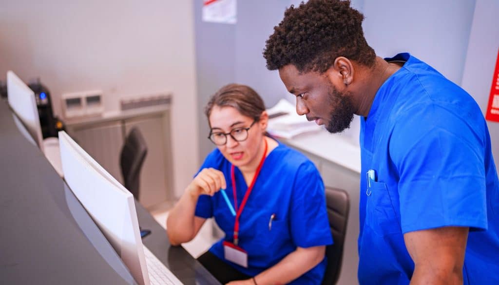 Two healthcare workers in blue scrubs look at a computer screen together.