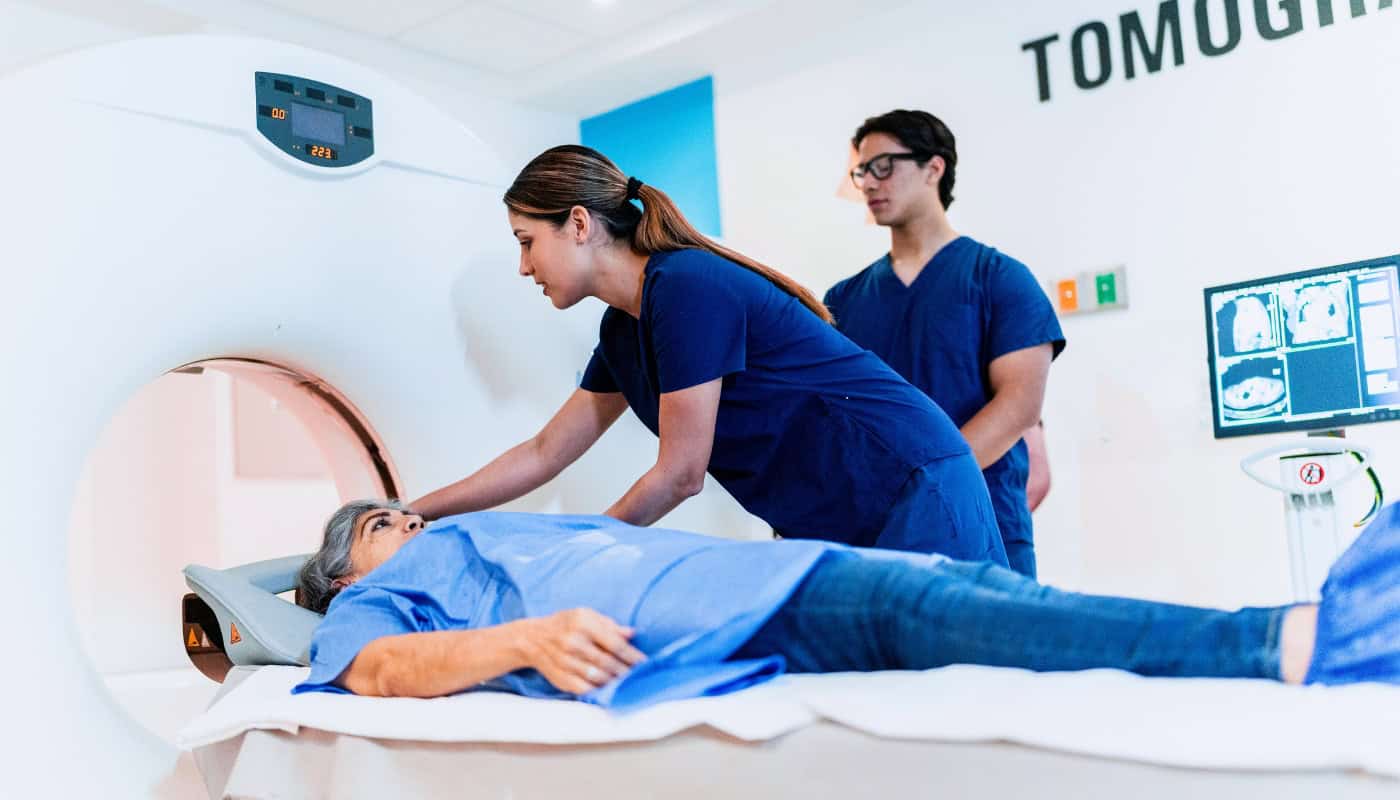A medical technician positions a patient lying on the table of a CT scanner while a colleague observes.