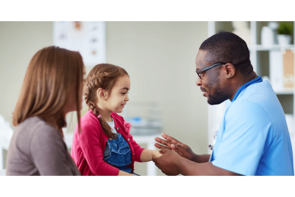 A doctor examining a young girl’s wrist while her mother sits beside her.